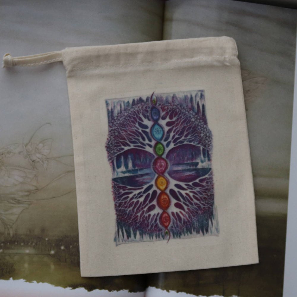 A small cream-colored fabric drawstring pouch featuring a colorful tree of life chakra print, displayed on an open book with printed pages visible.