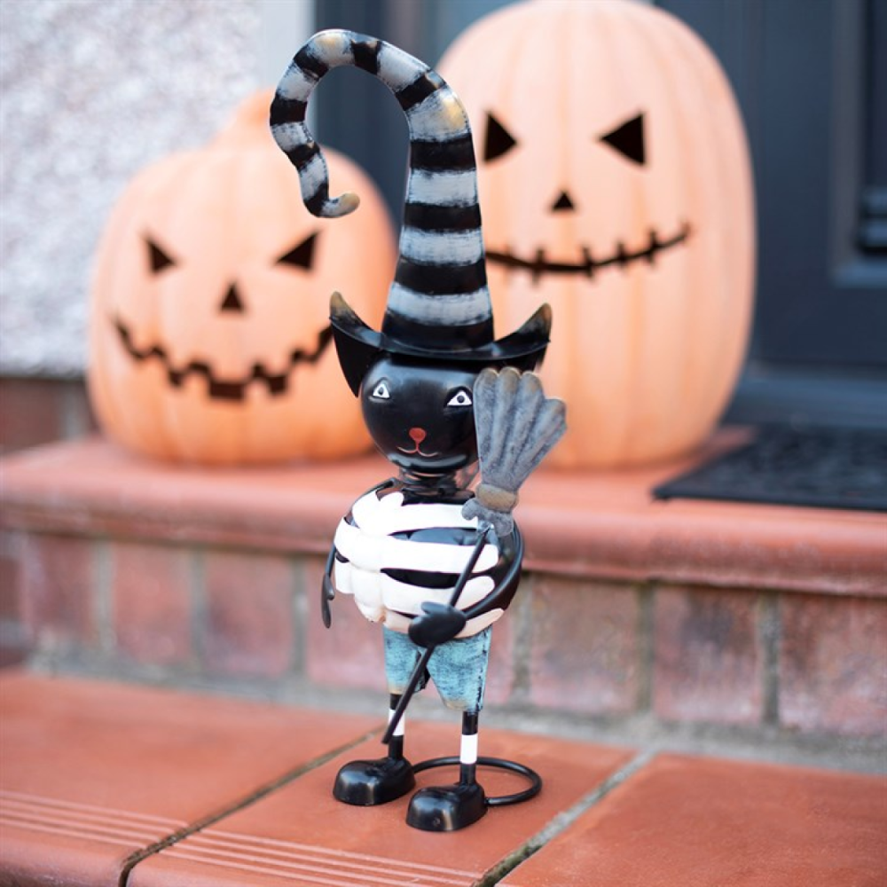 A Halloween decorative figurine of a black cat wrapped in a white bandage and wearing a striped black-and-white hat, holding a broom, displayed on a brick step with jack-o'-lanterns in the background.