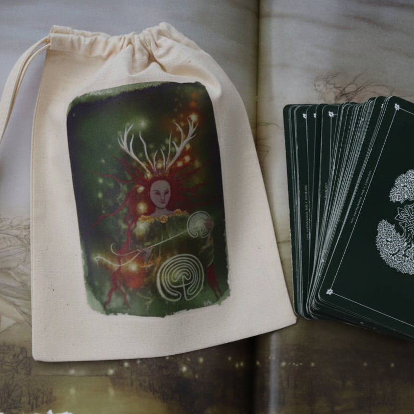 A cream-colored cotton drawstring pouch with a colorful fantasy illustration of a skull-headed figure with antlers, shown next to a fanned deck of dark green cards with silver circular symbols, laid out on an open book.