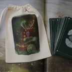 A cream-colored cotton drawstring pouch with a colorful fantasy illustration of a skull-headed figure with antlers, shown next to a fanned deck of dark green cards with silver circular symbols, laid out on an open book.