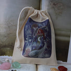 A beige fabric drawstring pouch with a colorful fantasy mermaid illustration, displayed on top of open books with crystals and a beaded necklace around it.