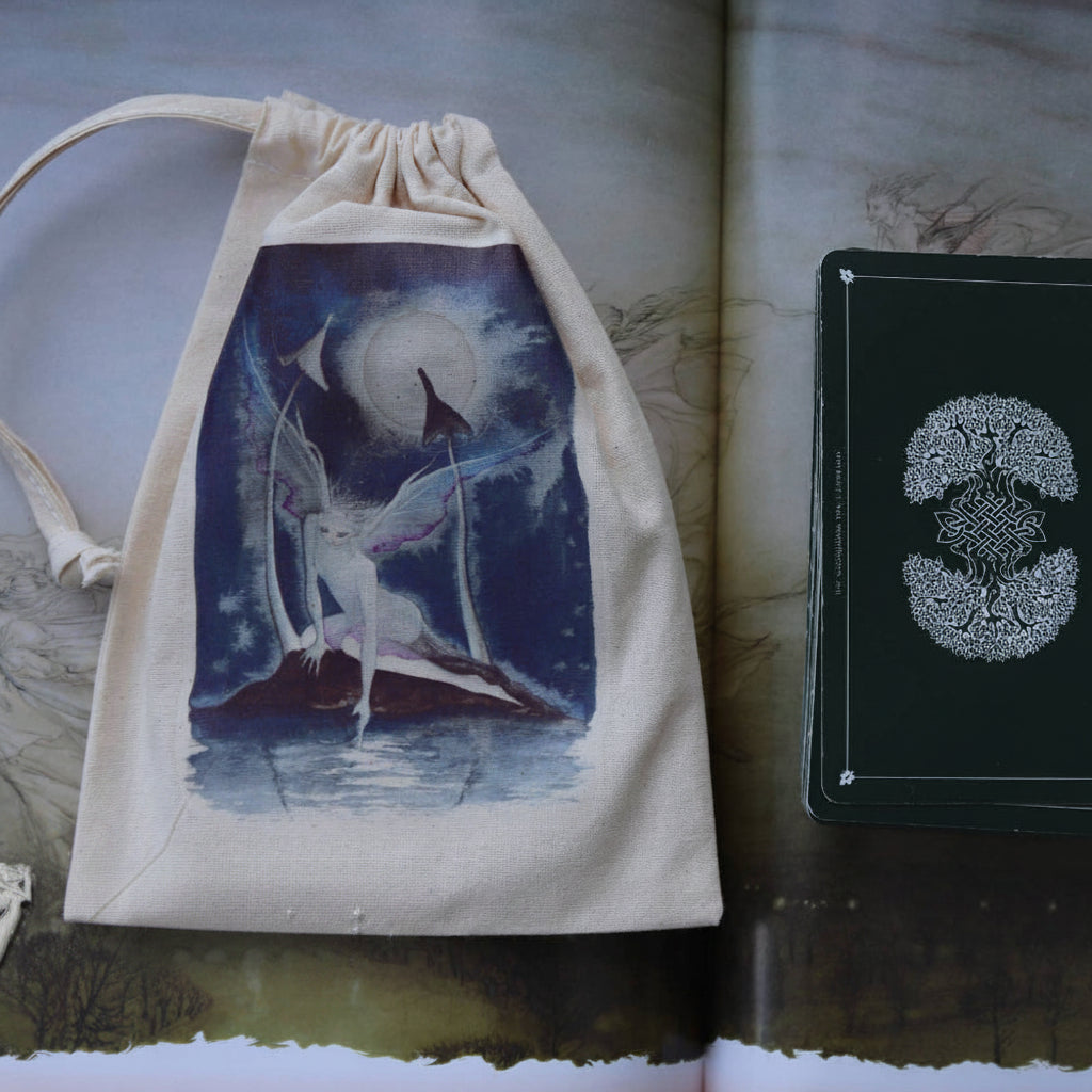 A cream fabric drawstring pouch printed with a blue and white fairy illustration, displayed next to a dark green deck box with a silver winged emblem, set against an open illustrated book.