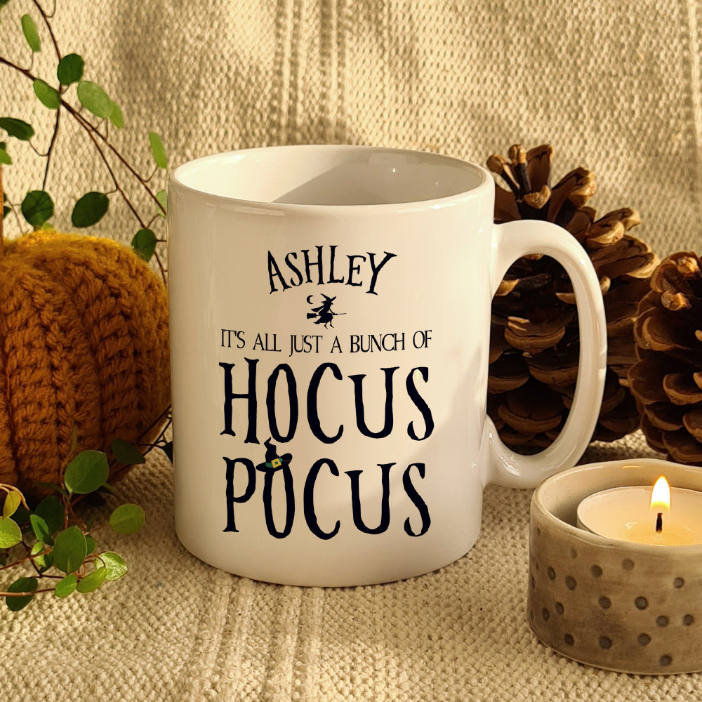 A white ceramic mug with 'ASHLEY' personalized text and 'It's all just a bunch of HOCUS POCUS' printed in black. The mug is displayed on a cozy background with pine cones and a candle.