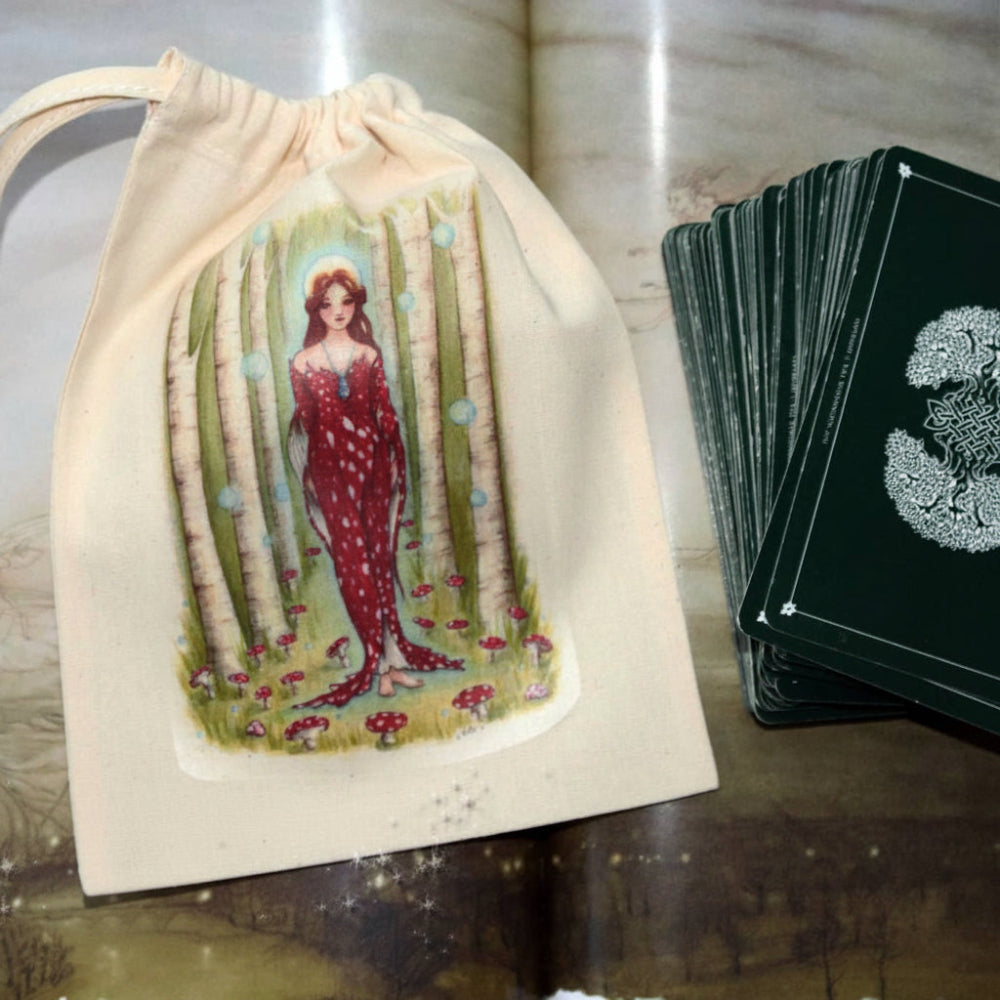 A cream fabric drawstring pouch printed with an illustration of a woman in a red dress among birch trees, shown next to a deck of dark green tarot cards on a rustic wooden surface.
