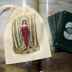 A cream fabric drawstring pouch printed with an illustration of a woman in a red dress among birch trees, shown next to a deck of dark green tarot cards on a rustic wooden surface.