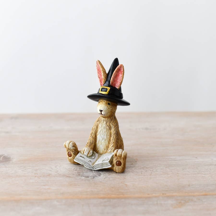 Figurine of a rabbit wearing a witch hat and reading a book on a wooden surface.