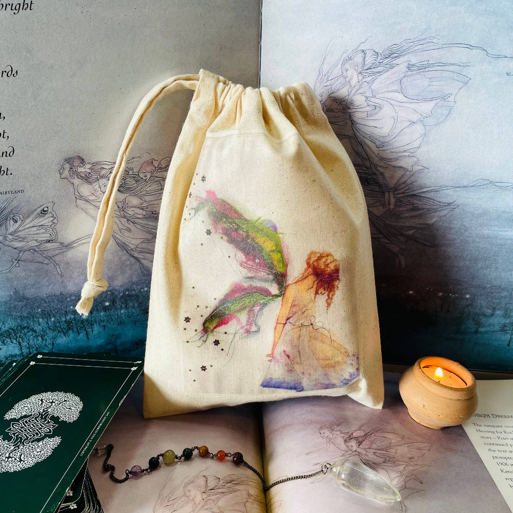 A cream-colored fabric drawstring pouch featuring a watercolor illustration of a fairy, styled on an open book with tarot cards, a candle, and crystals.