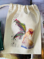 A cream-colored canvas drawstring pouch featuring a colorful watercolor illustration of a fairy with wings and a small figure, shown on a patterned surface.