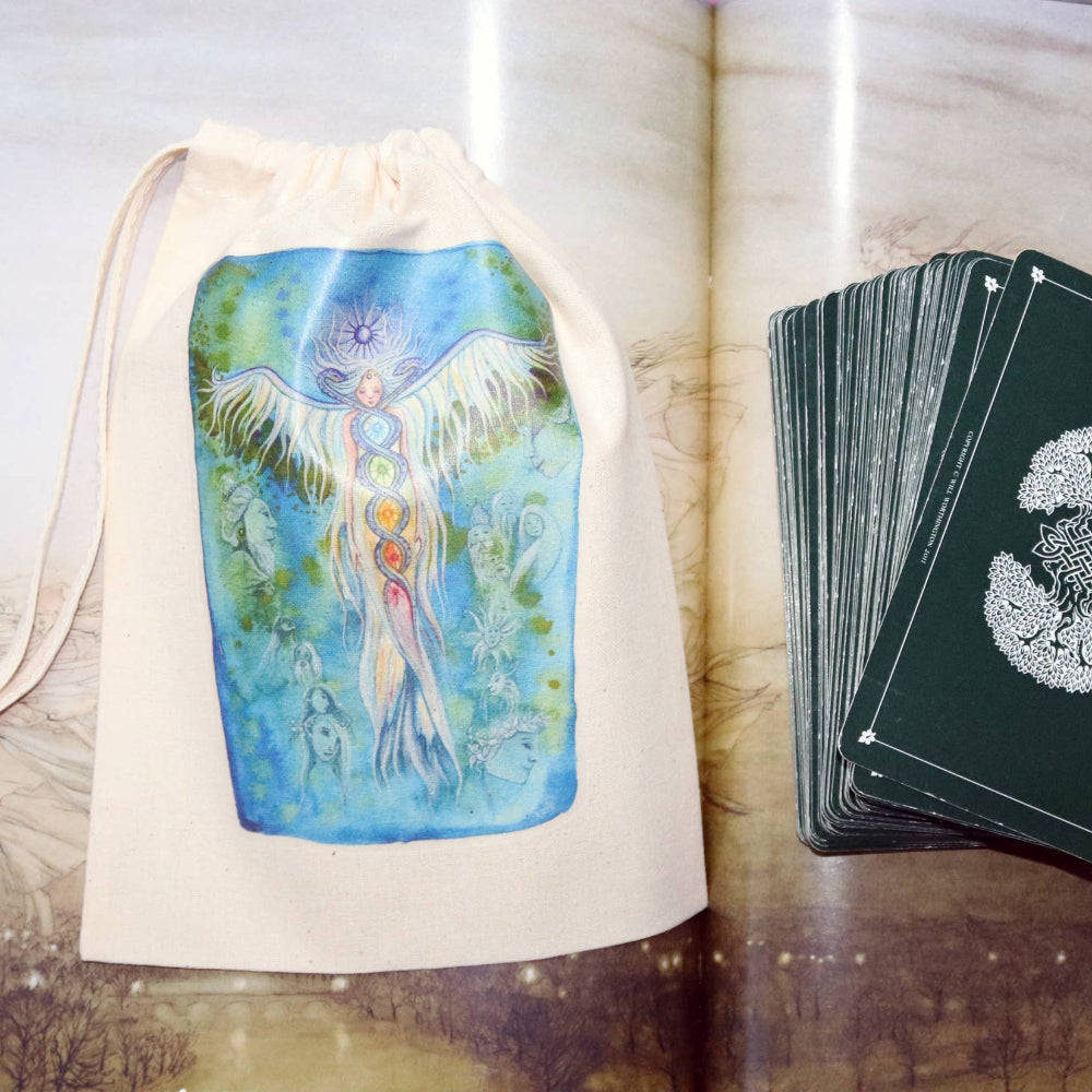 A tarot card deck with dark green cards featuring a white tree-of-life design, displayed next to a cream canvas drawstring bag printed with a colorful winged figure illustration, set against an open book with printed pages.