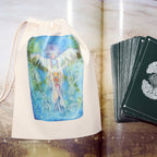 A tarot card deck with dark green cards featuring a white tree-of-life design, displayed next to a cream canvas drawstring bag printed with a colorful winged figure illustration, set against an open book with printed pages.