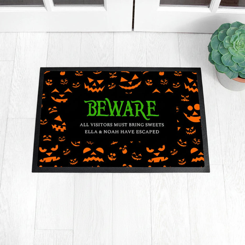 A rectangular black Halloween doormat with orange jack-o'-lantern faces, green 'BEWARE' lettering, and white text reading 'ALL VISITORS MUST BRING SWEETS ELLA & NOAH HAVE ESCAPED', shown on a white wood floor near a plant.