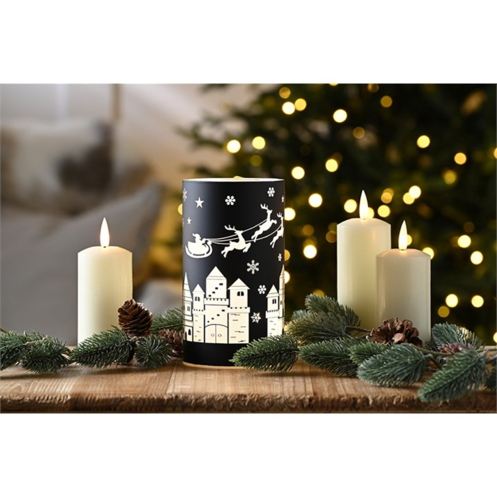A black cylindrical candle holder with a white Christmas village and Santa design, displayed on a wooden surface with lit white pillar candles, pine branches, and a blurred Christmas tree in the background.