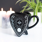 Black heart-shaped mug with mystical designs on a white surface with candles and a plant in the background