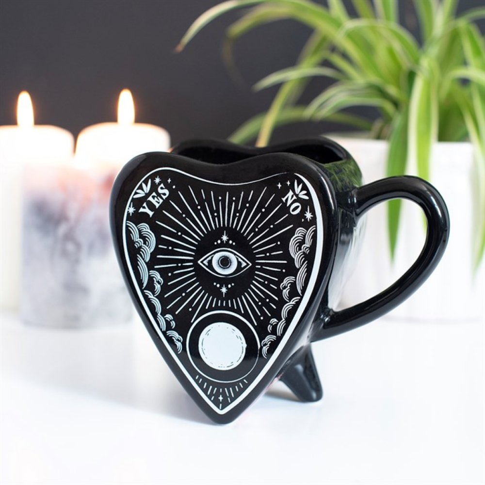 Black heart-shaped mug with mystical designs on a white surface with candles and a plant in the background