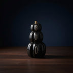 A glossy black stacked pumpkin decoration spelling 'BOOO' with white lettering, placed on a light wood surface against a neutral light gray background.
