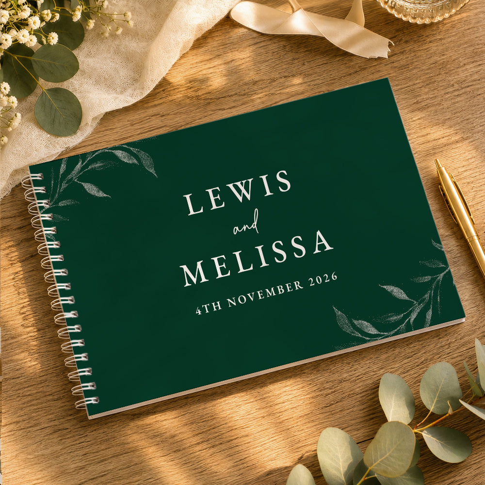 A personalized spiral-bound wedding guest book with a dark green cover, featuring the names 'LEWIS and MELISSA' and the date '4TH NOVEMBER 2026' in white text, styled on a wooden table with eucalyptus leaves and a gold pen.