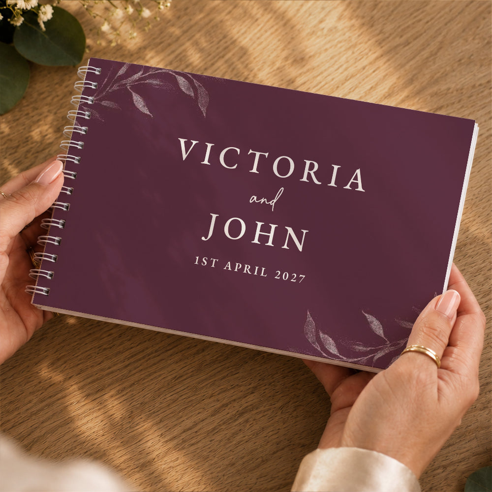 A personalized spiral-bound wedding guest book held by two hands over a wooden table. The cover is a deep plum purple with elegant white floral illustrations in the corners and the text 'VICTORIA and JOHN 1ST APRIL 2027' printed in white serif font.