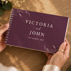 A personalized spiral-bound wedding guest book held by two hands over a wooden table. The cover is a deep plum purple with elegant white floral illustrations in the corners and the text 'VICTORIA and JOHN 1ST APRIL 2027' printed in white serif font.