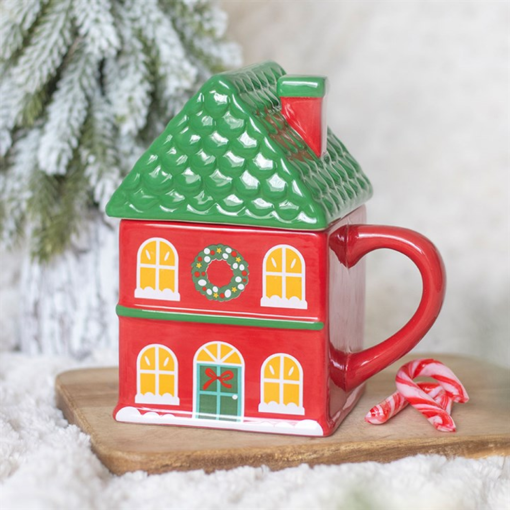 A ceramic mug shaped like a red house with a green scalloped roof, yellow window panes, a green wreath on the front, and a red handle. The mug sits on a wooden board with a candy cane and blurred evergreen branches in the background.