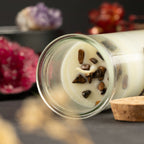 A clear glass jar candle lies on its side on a dark surface, showing creamy off-white wax with embedded dark brown stone chips and a white wick. A cork stopper rests beside the jar, with blurred decorative crystals in the background.