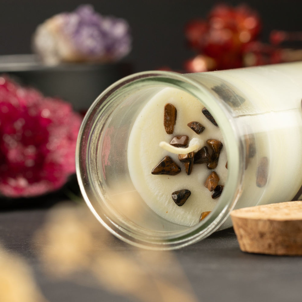 A clear glass jar candle lies on its side on a dark surface, showing creamy off-white wax with embedded dark brown stone chips and a white wick. A cork stopper rests beside the jar, with blurred decorative crystals in the background.