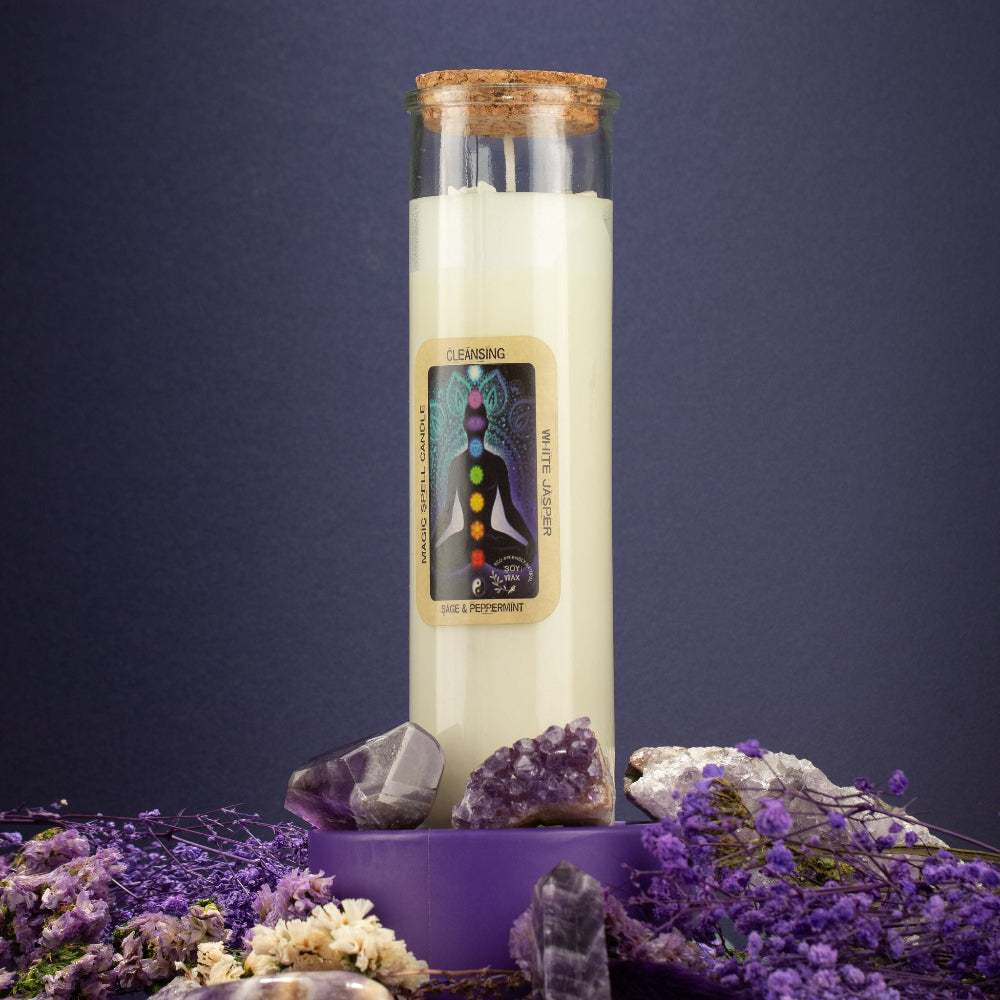 A glass jar candle with a cork stopper and cream-colored wax stands on a purple pedestal surrounded by purple crystals and dried lavender flowers against a dark blue background. The front label reads 'CLEANSING' and 'CHAKRA BALANCING'.