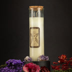 A tall glass jar candle with a cork stopper and cream-colored wax, featuring a brown label with a hand illustration. The candle is displayed on a black pedestal with purple and red dried flowers against a dark background.