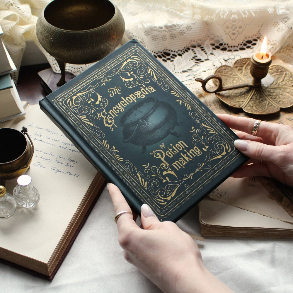 Vintage-style book titled 'The Encyclopedia of Potion Making' held by hands on a table with decorative items.