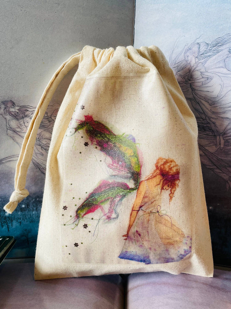 A cream-colored canvas drawstring bag featuring a colorful watercolor illustration of a fairy, set against a background with a gray wall and a printed sheet.
