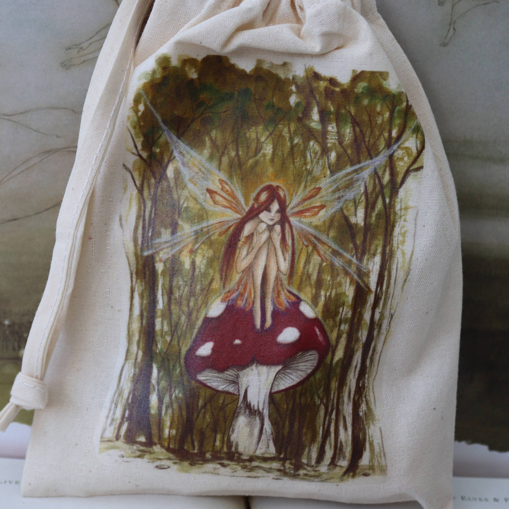 A cream-colored fabric drawstring pouch featuring a detailed illustration of a fairy sitting on a red and white mushroom in a forest, displayed on top of open vintage books.