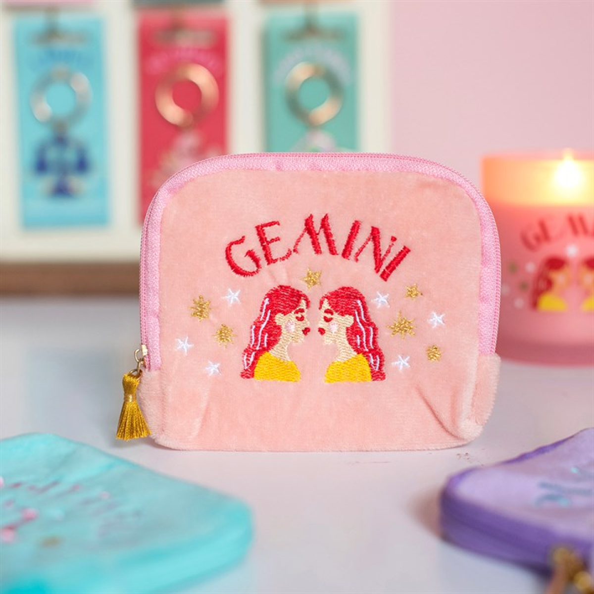 A pink velvet zippered pouch with 'GEMINI' embroidered in red and two Gemini zodiac figures with red hair, yellow tops, and gold stars. The pouch is shown on a tabletop with other colorful pouches blurred in the foreground and background, alongside a lit pink candle.