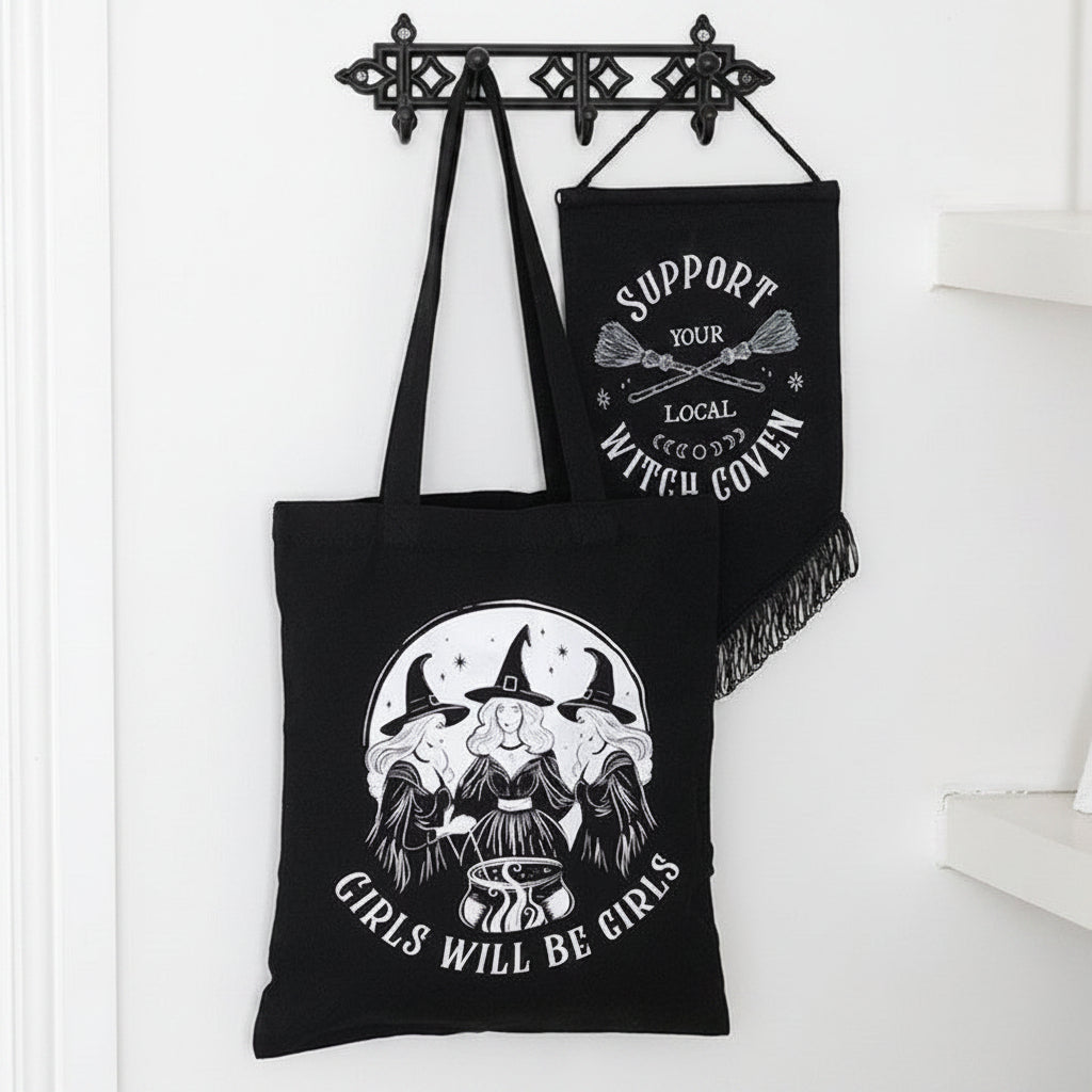 Black tote bag with witch-themed design and text, hanging on a wooden hook.