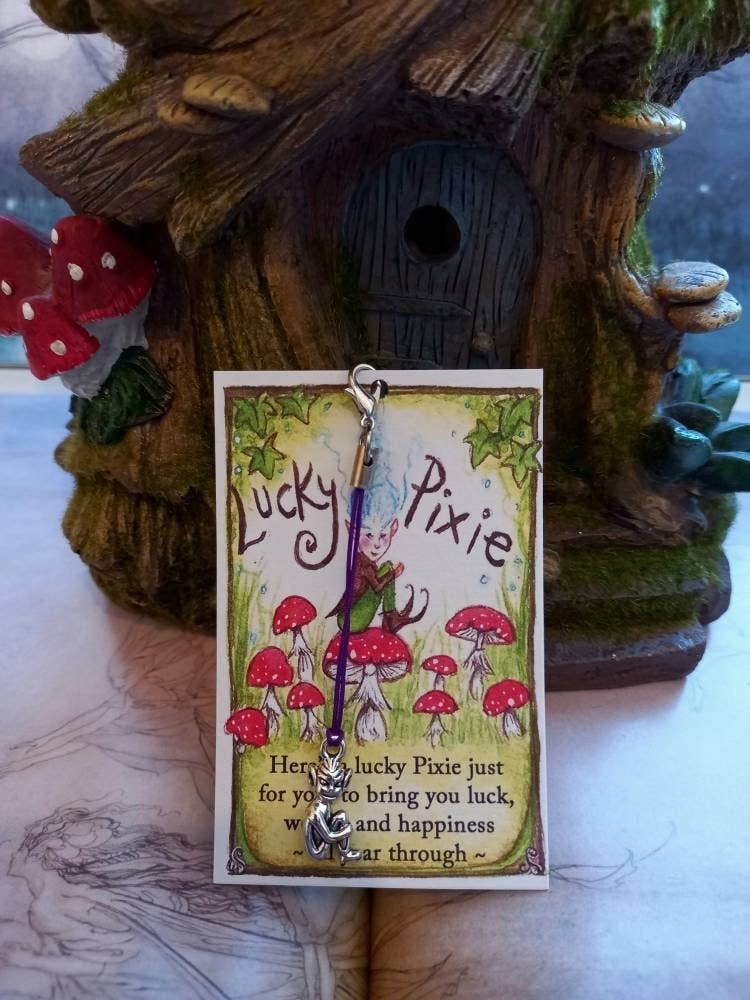 A Lucky Pixie charm with a purple string and silver cat pendant, displayed on its illustrated backing card in front of a decorative treehouse figurine.