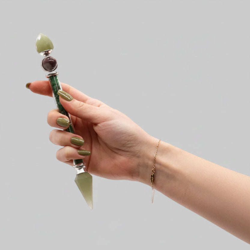 A hand holding a green crystal pointer wand with a pointed green stone tip, silver metal accents, and a dark purple gemstone, set against a solid gray background.