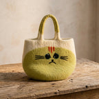 A cream and light yellow felt tote bag with a cute sad cat face design, styled on a beige stone surface next to a vase and books.