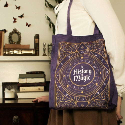 Person holding a purple tote bag with 'A History of Magic' design in a room with books and decor.