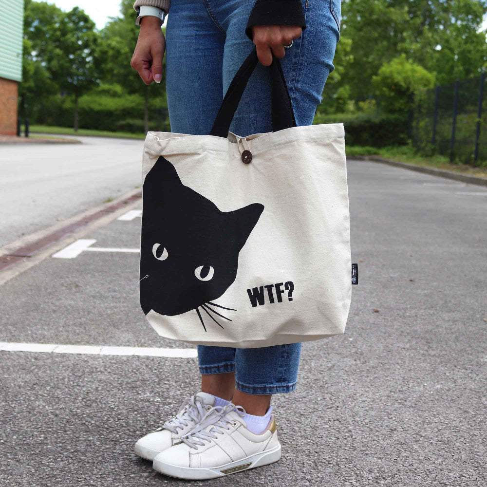 Person holding a tote bag with a black cat design and 'WTF?' text on a street.