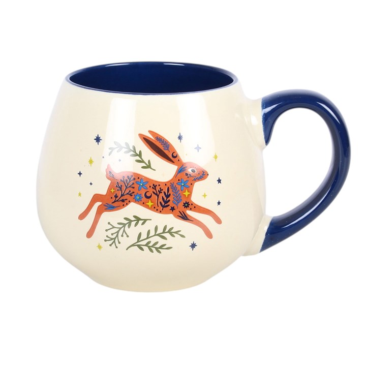 A cream-colored ceramic mug with a dark blue handle, interior, and exterior accents. The front of the mug features a colorful illustration of a jumping hare decorated with floral patterns, surrounded by small green leaves and yellow stars.