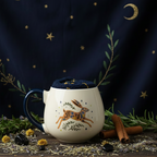 A cream-colored ceramic mug with a navy blue handle and interior, decorated with a colorful patterned rabbit and botanical motifs. The mug is filled with a navy blue textured fabric liner and sits on a round wooden tray alongside cinnamon sticks, with a blurred green plant in the background.