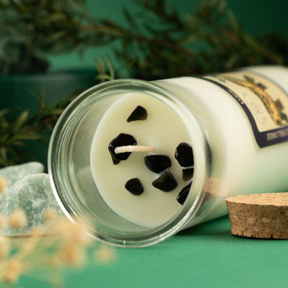 A clear glass jar candle lying on its side on a teal surface, with white wax, black stone-like accents, a visible wick, a cork stopper, and a decorative label. Greenery and crystals are softly blurred in the background.