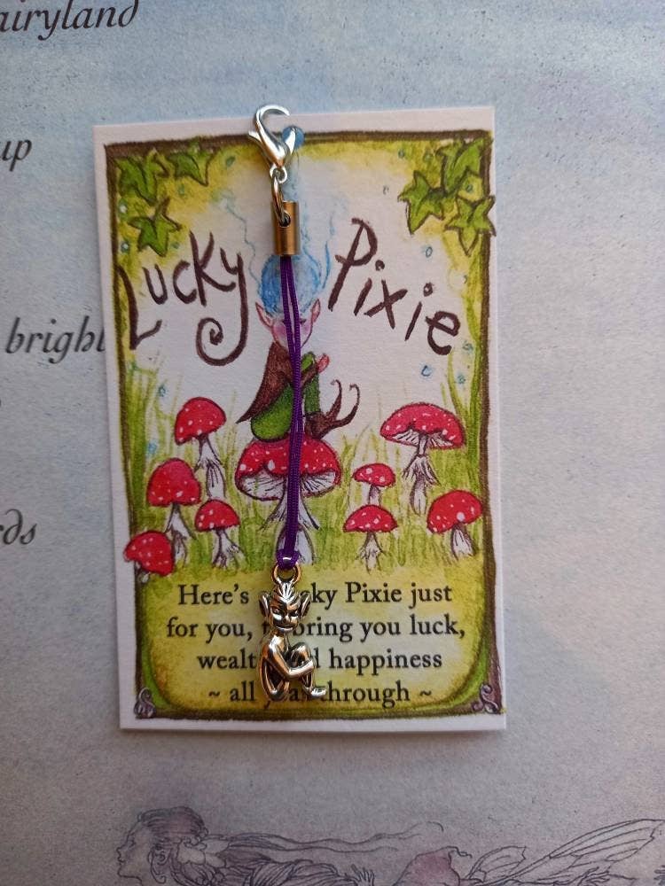 A Lucky Pixie charm with a purple string and silver-toned metal clasp and dangling letter charm, attached to a decorative card featuring red mushrooms and the words 'Lucky Pixie' on a light background.