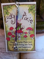 A Lucky Pixie charm with a silver heart and purple string, hanging from a card that features an illustration of a pixie among red mushrooms and the text 'Lucky Pixie'.