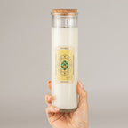 A hand holding a tall glass jar candle with a cork stopper. The candle wax is off-white, and the front label reads 'HAPPINESS' with a green lotus design
