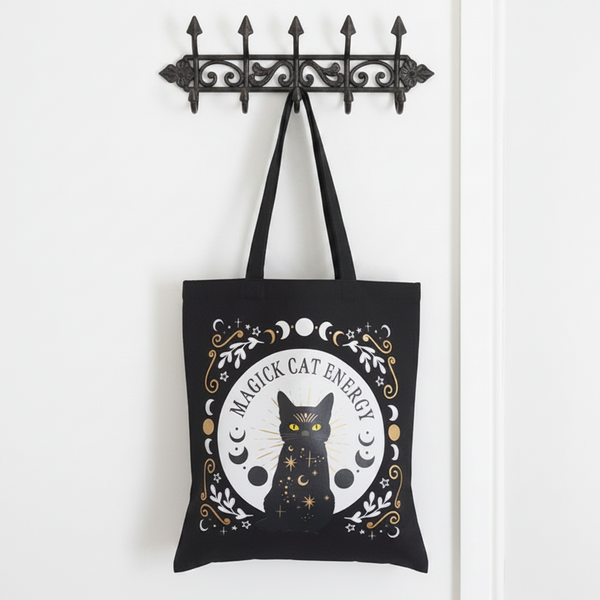 Black tote bag with a cat design and 'Magick Cat Energy' text hanging on a metal hook.