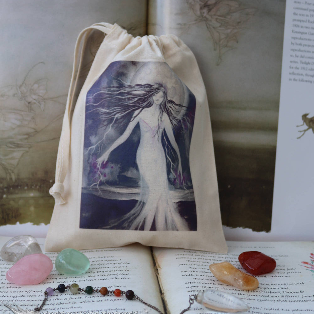 A beige fabric drawstring pouch featuring a printed illustration of a white fairy-like figure against a dark purple background, displayed on top of open books with crystals and a beaded necklace around it.