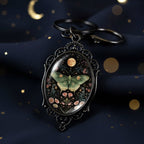 An ornate oval keychain pendant with a black filigree frame, featuring a moth and floral design under a moon on a dark background, resting on a white stone.