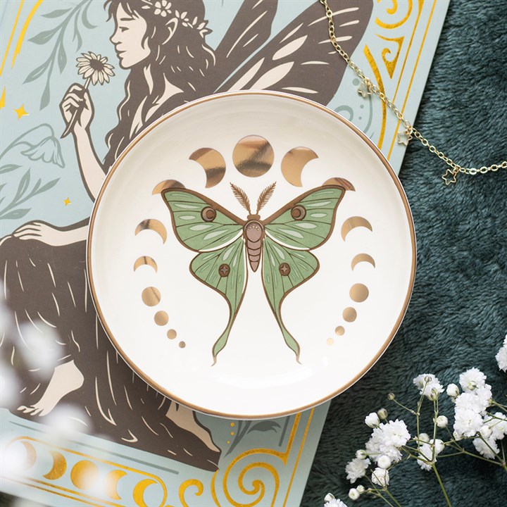 A white ceramic bowl with a gold rim, featuring a green luna moth and gold moon phase illustration, styled on top of a fairy art print with a gold chain necklace and baby's breath flowers.