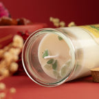 A glass jar candle with creamy off-white wax and green botanical pieces inside, shown lying on its side on a red surface with dried flowers and a red background.