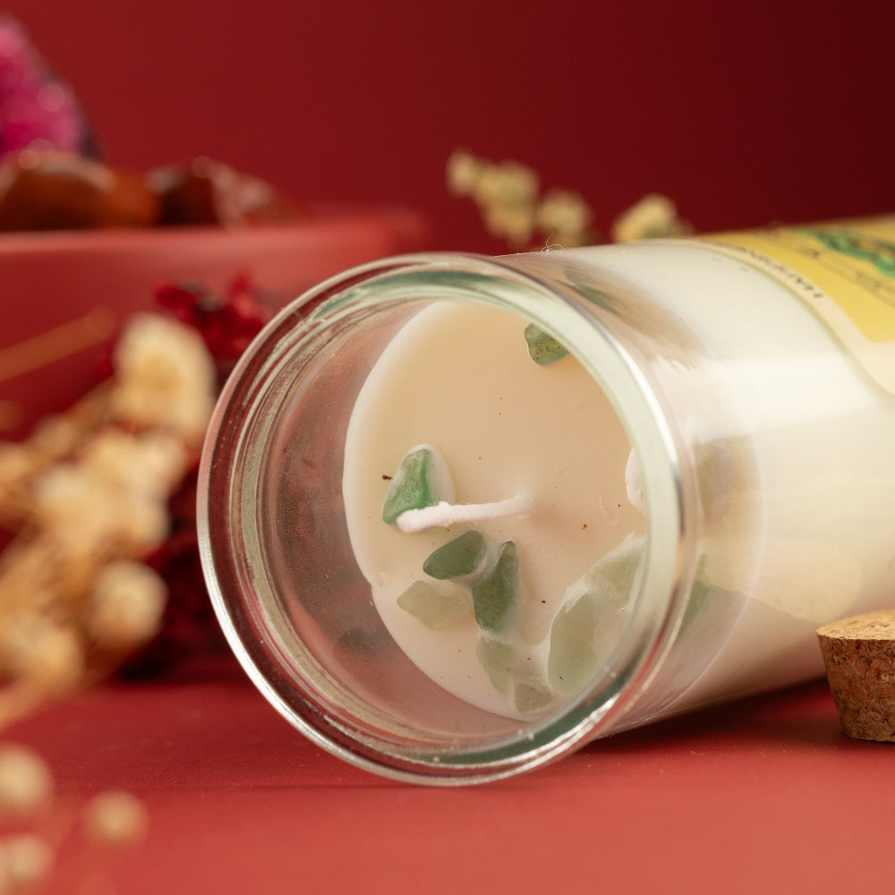 A glass jar candle with creamy off-white wax and green botanical pieces inside, shown lying on its side on a red surface with dried flowers and a red background.