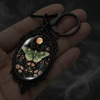 A close-up of a hand holding a black metal keychain with an ornate oval pendant. The pendant features a dark fantasy-style illustration of a green butterfly surrounded by small flowers under a moonlit sky with a moon and sun.